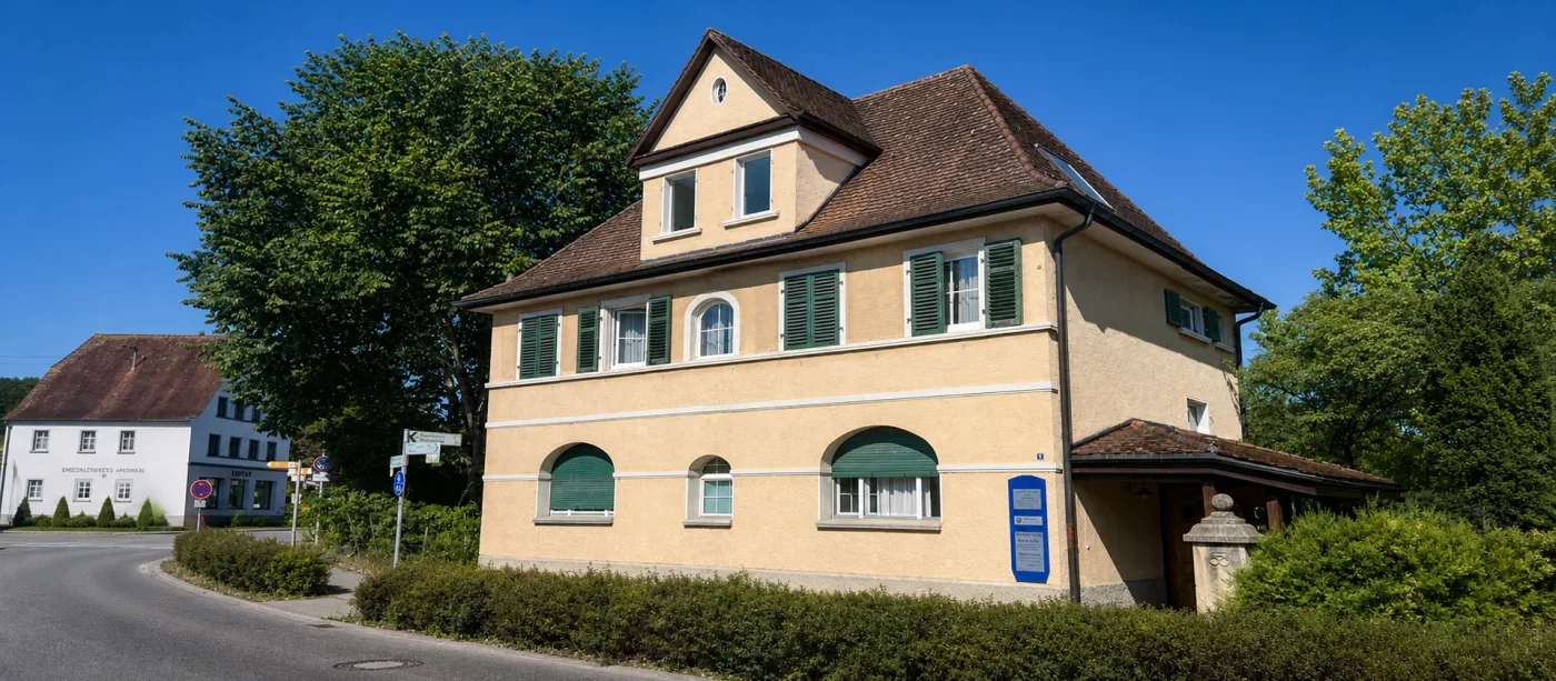 Das Praxishaus von der Straße aus: gelber Putzbau mit rotem Ziegeldach und Hecke davor.